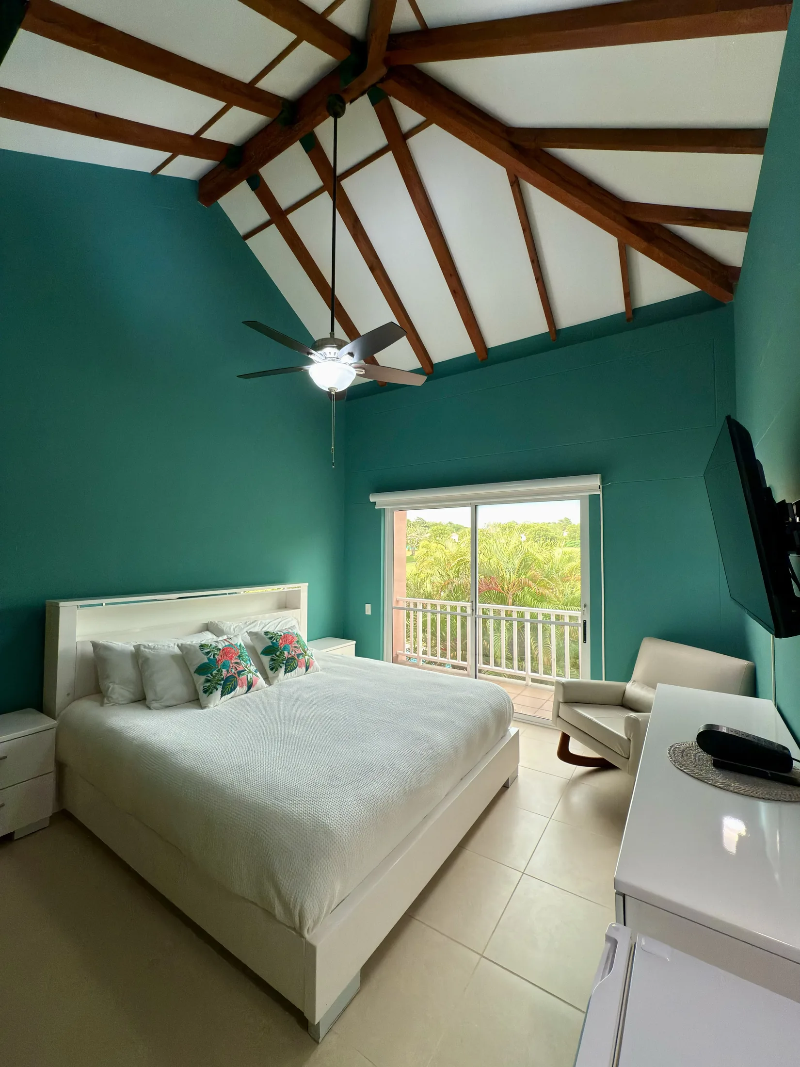 Teal bedroom with vaulted wood-beam ceiling, ceiling fan, balcony door, and white furniture
