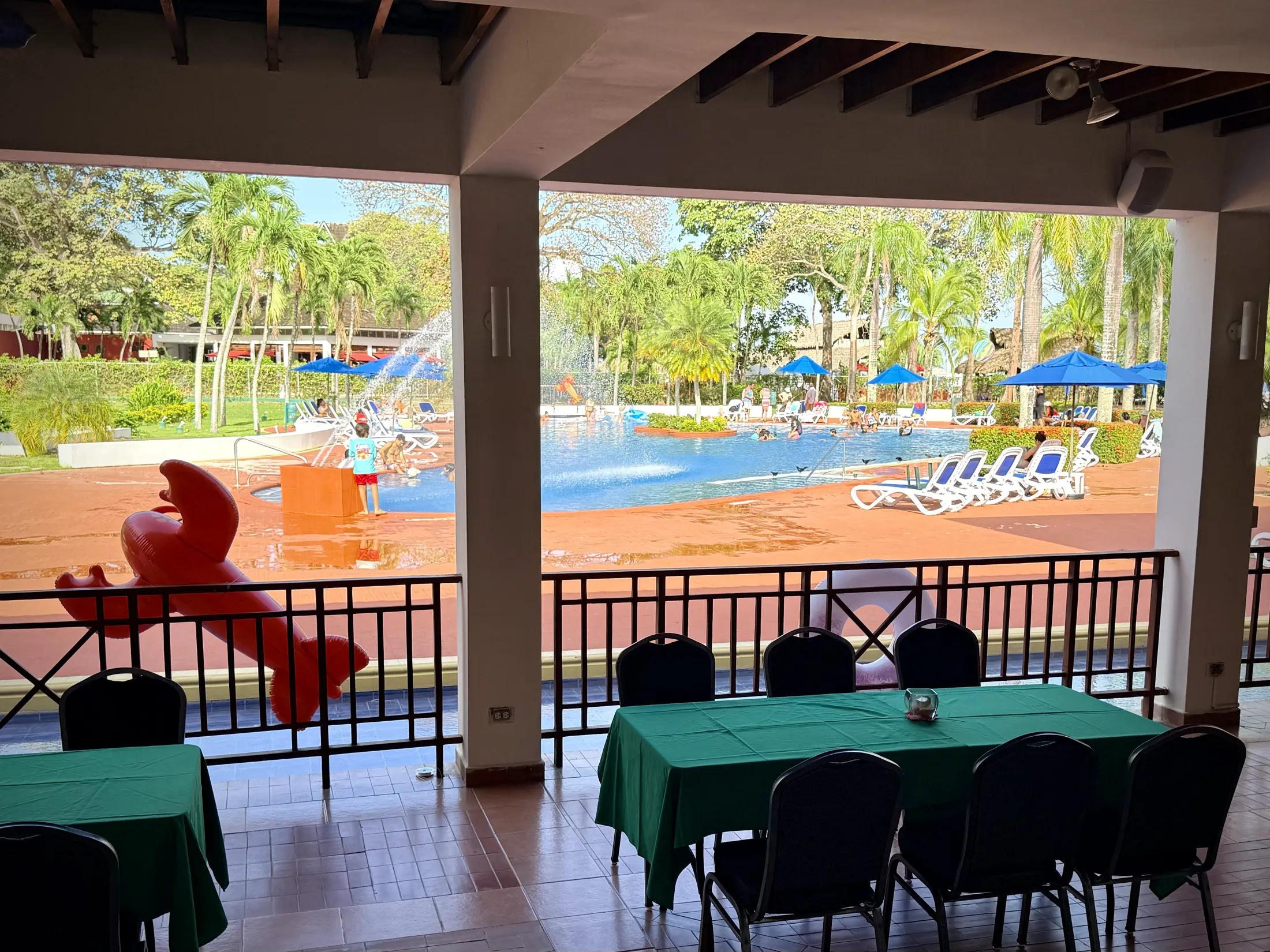 Resort pool area with fountains, lounge chairs, blue umbrellas, and covered dining terrace