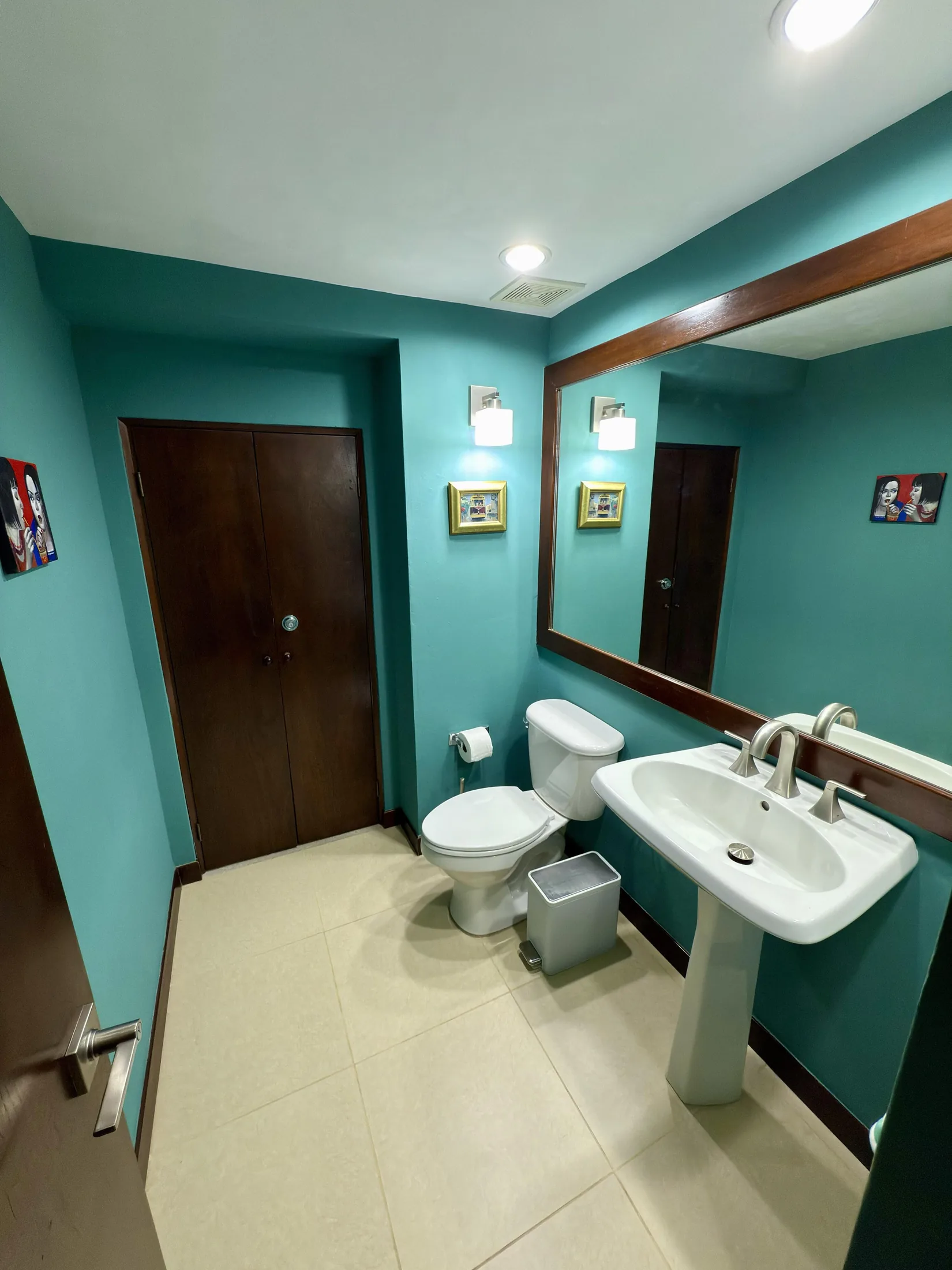 Teal-walled bathroom with pedestal sink, toilet, large wood-framed mirror, and art