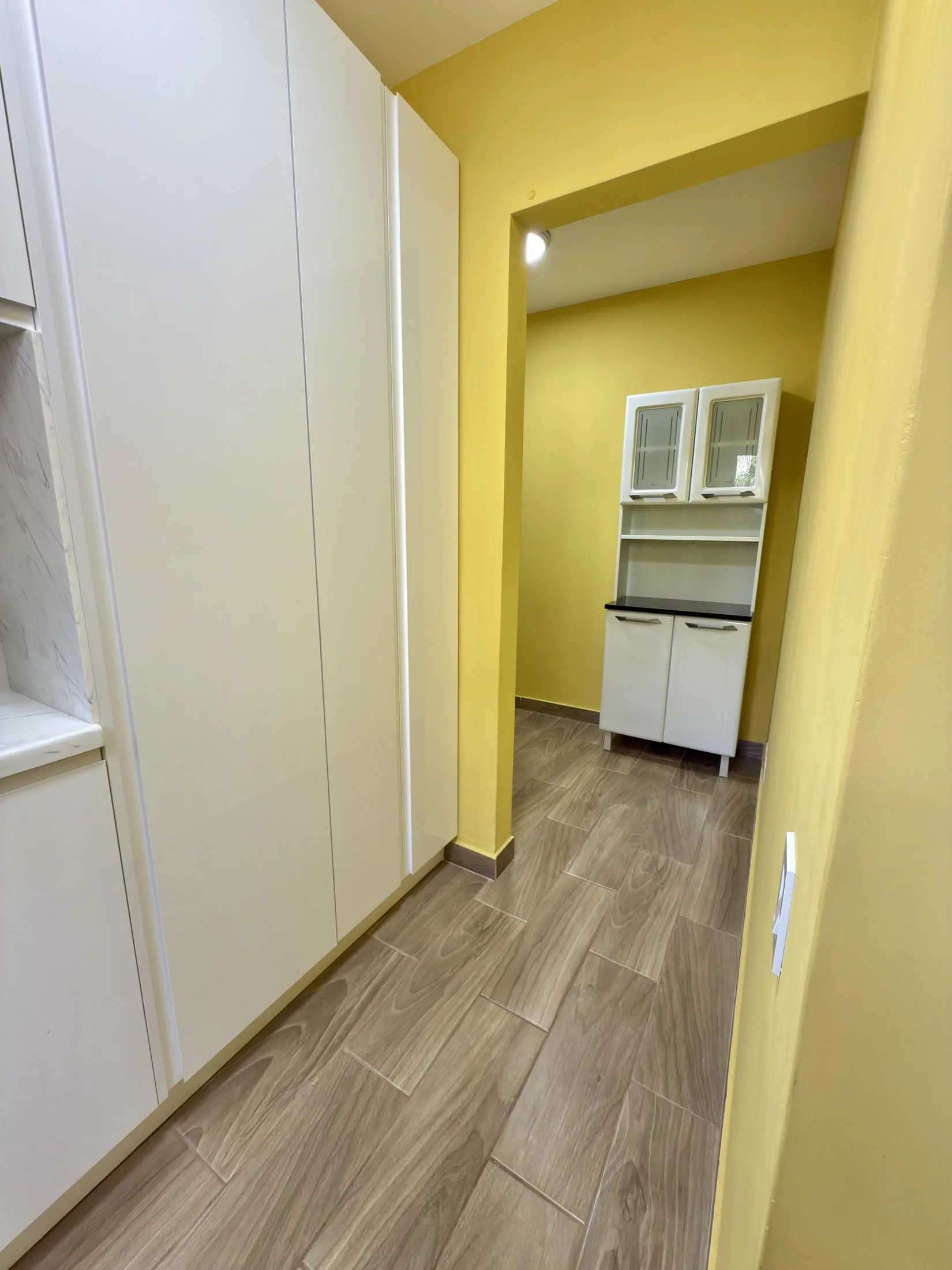 Pantry corridor with yellow walls, tall white cabinets, and glass-front upper cabinet