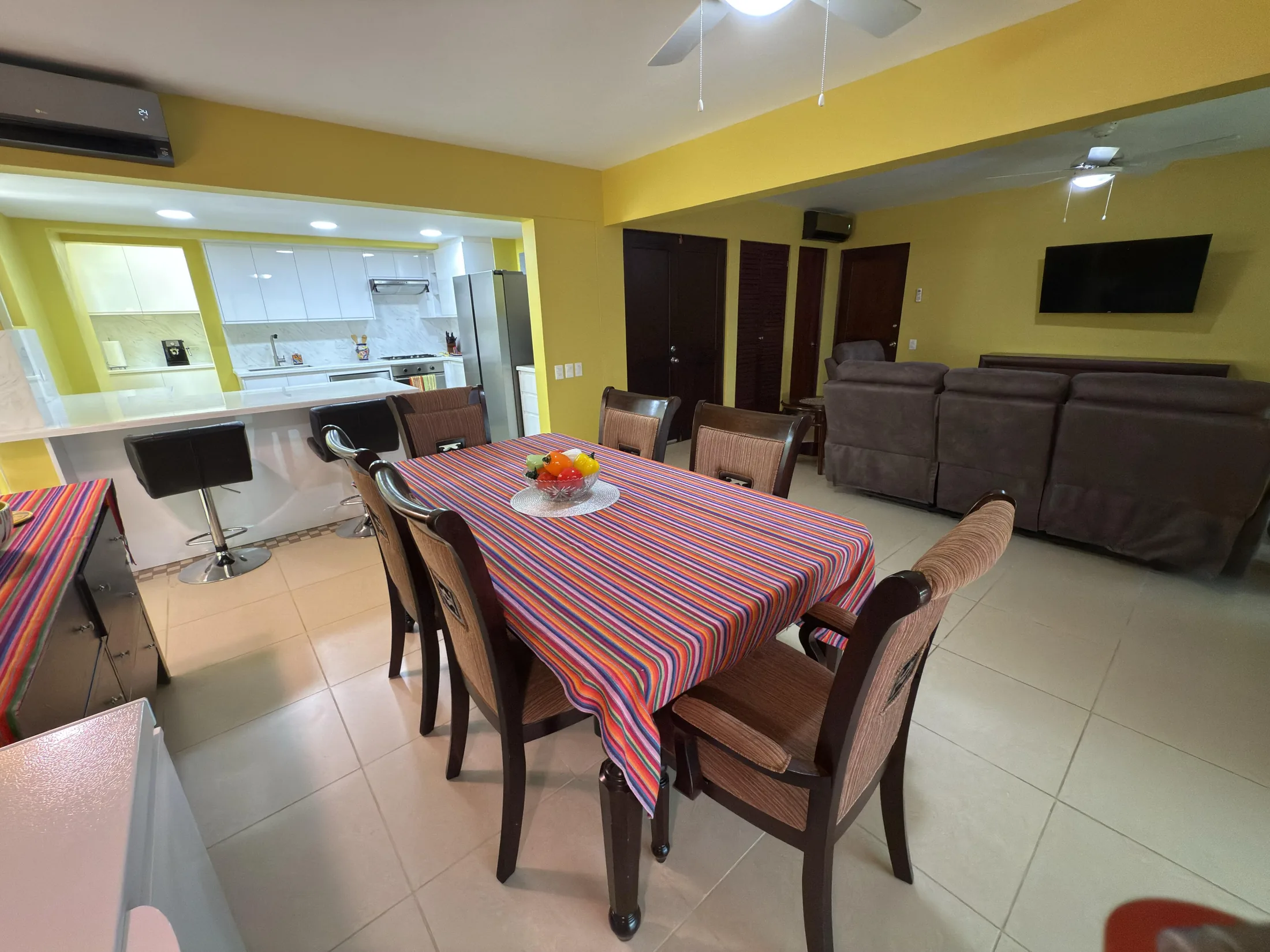Wide shot of open-plan dining table with striped cloth, kitchen bar with stools, and lounge area